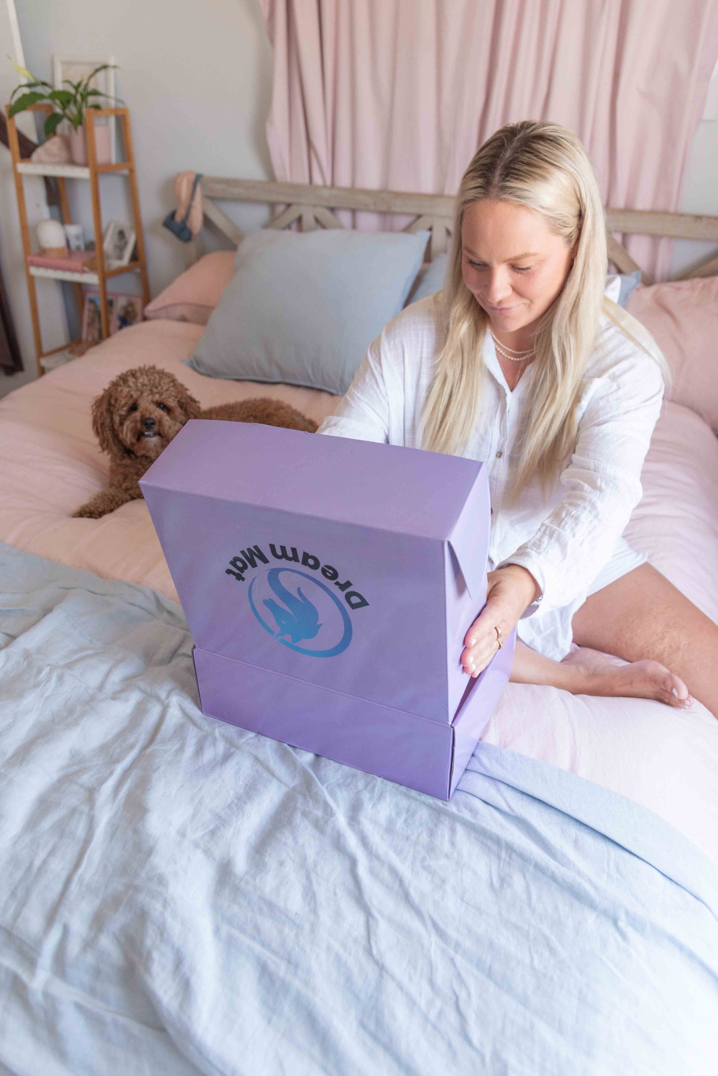 Woman opening a purple box with 'Dream Matt' branding on a bed with a dog beside her.